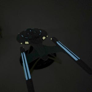 Puede incluir: Un modelo de nave espacial negro y azul con una parte superior circular y dos alas largas y delgadas. Las alas tienen una franja azul en el centro.