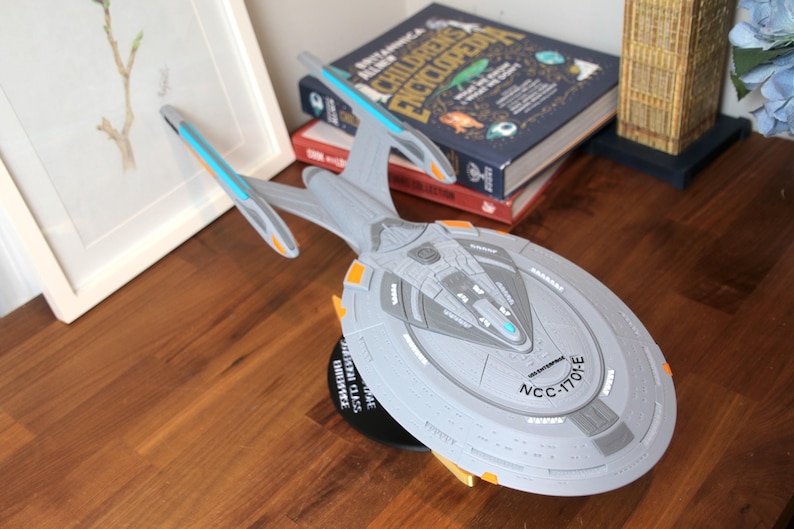 Puede incluir: Un modelo gris y azul de la nave espacial USS Enterprise NCC-1701-E de Star Trek. El modelo est&aacute; sobre una base negra con un borde dorado. La nave es un objeto de colecci&oacute;n popular para los fans de la franquicia Star Trek.