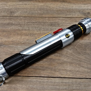 Puede incluir: Un accesorio de sable de luz plateado y negro con un bot&oacute;n rojo y detalles dorados. El accesorio tiene un dise&ntilde;o cil&iacute;ndrico con un anillo en la base. El sable de luz est&aacute; sobre una superficie de madera.