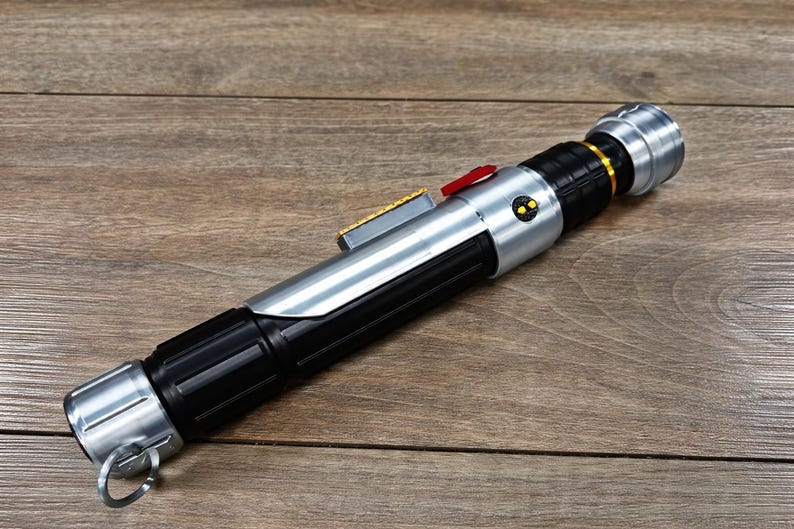 Puede incluir: Un accesorio de sable de luz plateado y negro con un bot&oacute;n rojo y detalles dorados. El accesorio tiene un dise&ntilde;o cil&iacute;ndrico con un anillo en la base. El sable de luz est&aacute; sobre una superficie de madera.