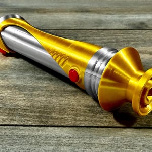 May include: A gold and silver lightsaber prop with red buttons. The hilt is gold with a silver centre section. The lightsaber is lying on a wooden surface.
