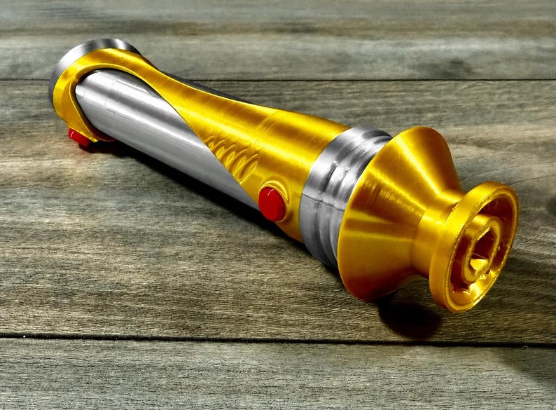 May include: A gold and silver lightsaber prop with red buttons. The hilt is gold with a silver center section. The lightsaber is lying on a wooden surface.