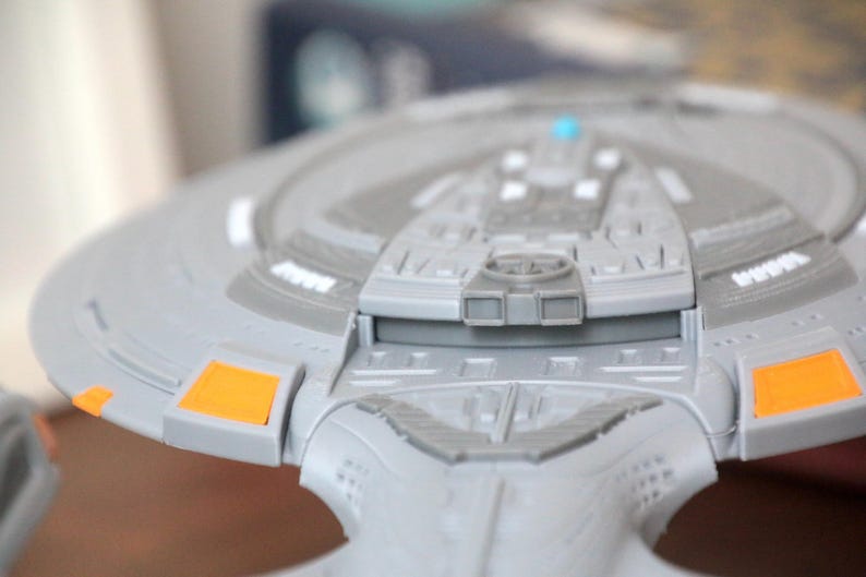 Puede incluir: Un modelo gris y naranja de una nave espacial con un dise&ntilde;o detallado. El modelo tiene una secci&oacute;n de puente elevada con una peque&ntilde;a luz azul.