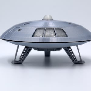 Puede incluir: Modelo gris de platillo volante con cúpula y tres patas de aterrizaje. El platillo tiene ventanas rectangulares y una pequeña cúpula transparente. Un artículo de colección o decoración.