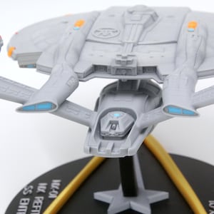 Puede incluir: Maqueta gris de una nave espacial con detalles en naranja y azul, montada sobre una base negra con soportes dorados. El modelo presenta alas detalladas y un cuerpo central, que se asemeja a una nave espacial de ciencia ficci&oacute;n.