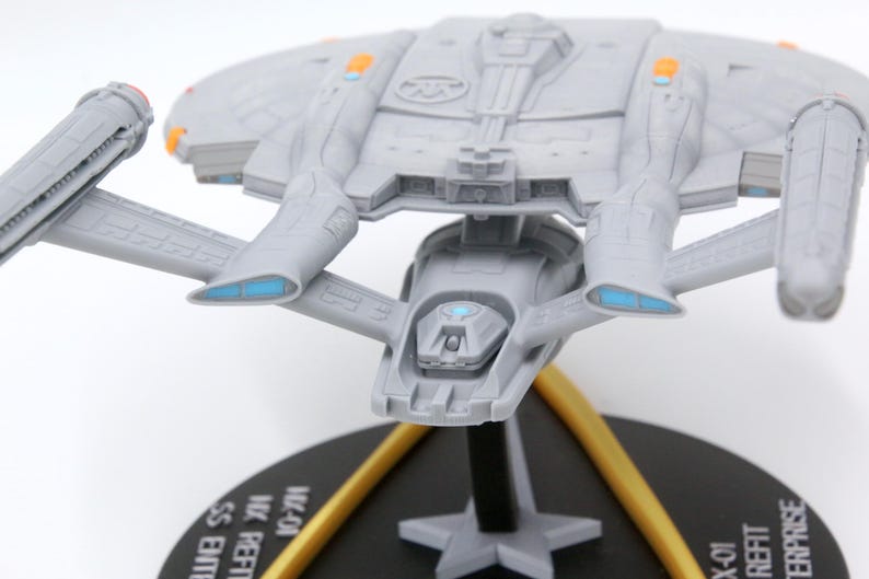 Puede incluir: Maqueta gris de una nave espacial con detalles en naranja y azul, montada sobre una base negra con soportes dorados. El modelo presenta alas detalladas y un cuerpo central, que se asemeja a una nave espacial de ciencia ficci&oacute;n.