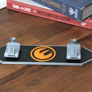 Puede incluir: Un expositor rectangular negro y gris con dos modelos de veh&iacute;culos grises y un emblema naranja. El expositor est&aacute; sobre una superficie de madera, con libros en el fondo.
