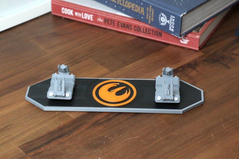 Puede incluir: Un expositor rectangular negro y gris con dos modelos de veh&iacute;culos grises y un emblema naranja. El expositor est&aacute; sobre una superficie de madera, con libros en el fondo.