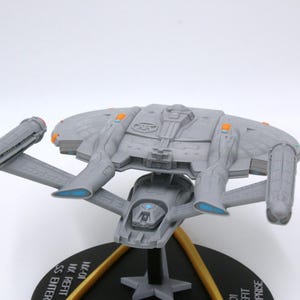 Puede incluir: Maqueta gris de una nave espacial con detalles intrincados, incluyendo alas y una estructura central. Tiene detalles en naranja y azul y est&aacute; montada sobre una base negra con soportes dorados. La base tiene texto.