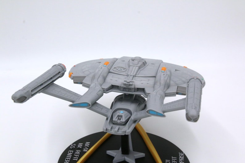 Puede incluir: Maqueta gris de una nave espacial con detalles intrincados, incluyendo alas y una estructura central. Tiene detalles en naranja y azul y est&aacute; montada sobre una base negra con soportes dorados. La base tiene texto.
