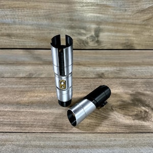 May include: Two silver and black lightsaber hilts are displayed on a wooden surface. One hilt is upright, while the other is lying on its side. The hilts have a metallic finish with gold-colored accents.