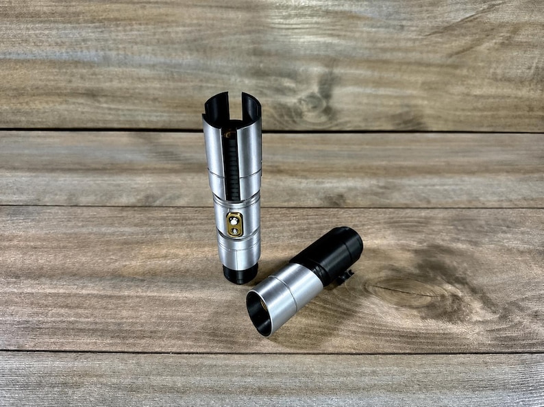 May include: Two silver and black lightsaber hilts are displayed on a wooden surface. One hilt is upright, while the other is lying on its side. The hilts have a metallic finish with gold-colored accents.