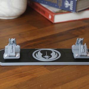 Może przedstawiać: Czarny i szary stojak na miecz świetlny Star Wars z dwoma szarymi uchwytami na miecze świetlne. Stojak posiada srebrny emblemat i jest prezentowany na drewnianej powierzchni.