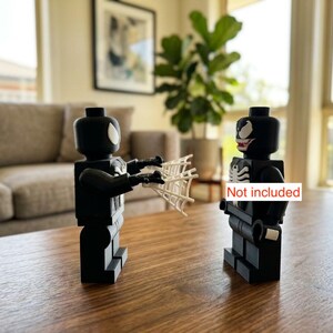 May include: Two black Lego figures, one holding a white web, facing each other. One figure has a white spider symbol, the other has a white spider symbol and a red mouth. The figures are on a brown table.