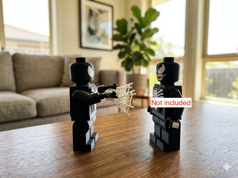 May include: Two black Lego figures, one holding a white web, facing each other. One figure has a white spider symbol, the other has a white spider symbol and a red mouth. The figures are on a brown table.