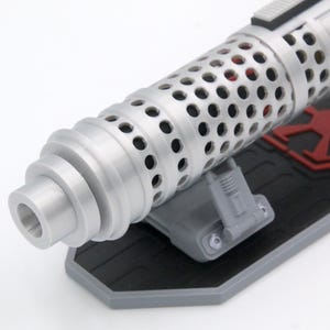 May include: A silver-colored cylindrical object with a perforated surface texture, mounted on a black rectangular base featuring a red geometric emblem. The object has a stepped, tapered end and is supported by a small gray bracket.