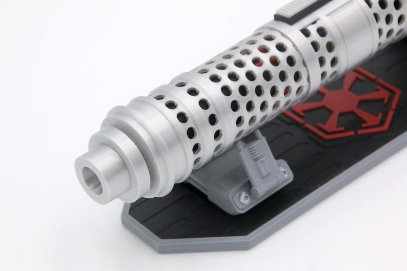 May include: A silver-colored cylindrical object with a perforated surface texture, mounted on a black rectangular base featuring a red geometric emblem. The object has a stepped, tapered end and is supported by a small gray bracket.