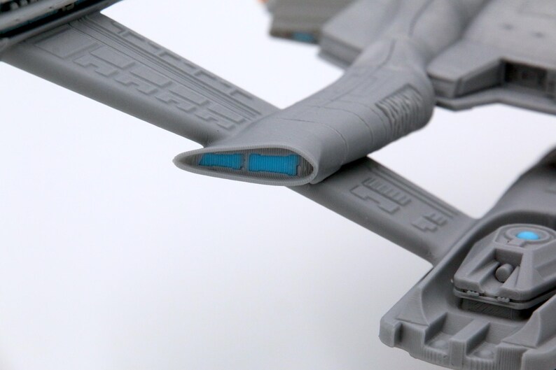 Puede incluir: Modelo gris detallado de una nave espacial, con paneles intrincados y detalles azules. El dise&ntilde;o incluye estructuras en forma de ala y un cuerpo central con un elemento azul brillante, lo que sugiere un tema futurista o de ciencia ficci&oacute;n.