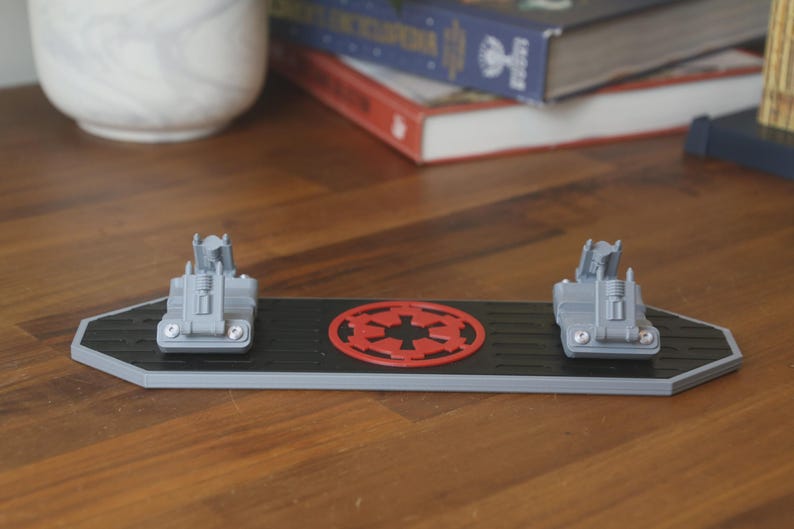 May include: A black and gray Star Wars-themed desk organizer with two gray vehicle models and a red Imperial symbol. The organizer is rectangular with angled sides and sits on a wooden surface.