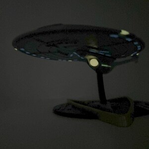 Puede incluir: Un modelo gris oscuro de una nave espacial con luces brillantes en la parte inferior. La nave espacial est&aacute; en un soporte negro.