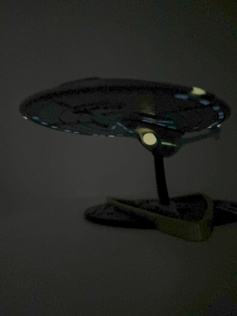 Puede incluir: Un modelo gris oscuro de una nave espacial con luces brillantes en la parte inferior. La nave espacial est&aacute; en un soporte negro.