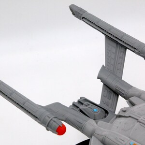 Puede incluir: Modelo gris de una nave espacial con dos largas g&oacute;ndolas de curvatura, cada una con una punta roja. La nave presenta un dise&ntilde;o detallado con acentos azules y naranjas, sobre un fondo blanco.