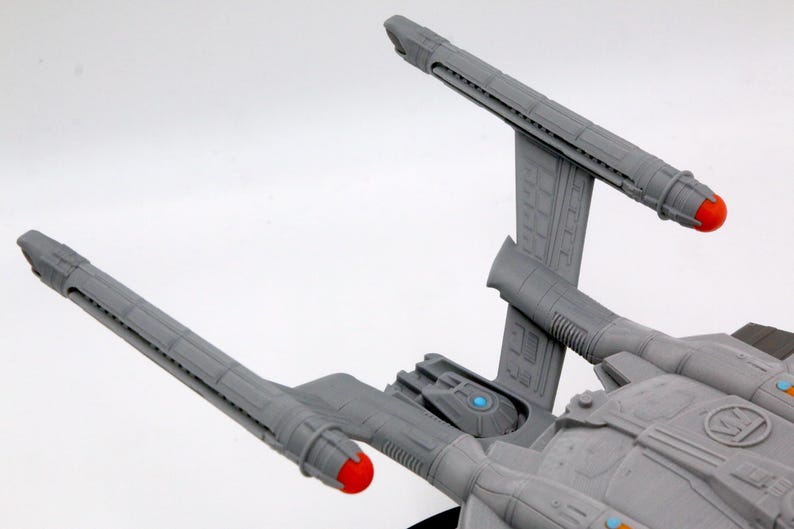 Puede incluir: Modelo gris de una nave espacial con dos largas g&oacute;ndolas de curvatura, cada una con una punta roja. La nave presenta un dise&ntilde;o detallado con acentos azules y naranjas, sobre un fondo blanco.