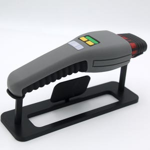 K&ouml;nnte beinhalten: Ein graues und rotes Handger&auml;t mit einem schwarzen St&auml;nder. Das Ger&auml;t hat einen strukturierten Griff und ein kleines Display mit gr&uuml;nen Lichtern.