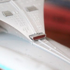 K&ouml;nnte beinhalten: Nahaufnahme eines wei&szlig;en Raumschiffmodells mit einem roten Licht an der Unterseite. Das Raumschiff hat ein detailliertes Design mit einer strukturierten Oberfl&auml;che.