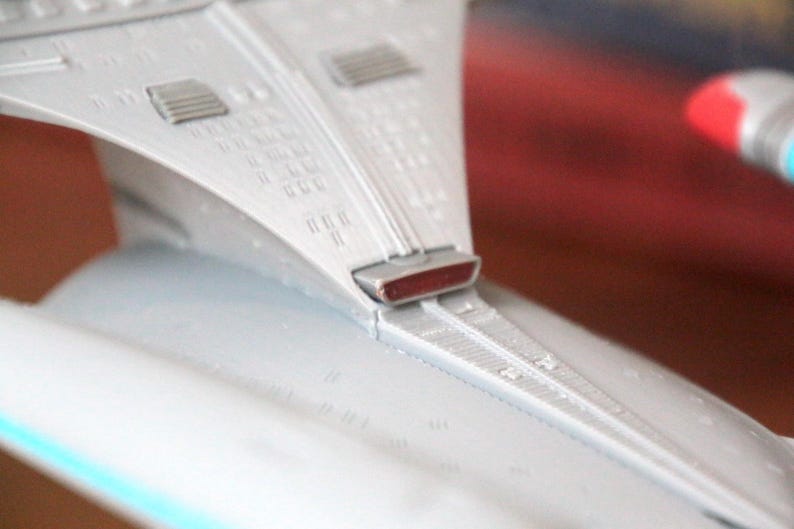 K&ouml;nnte beinhalten: Nahaufnahme eines wei&szlig;en Raumschiffmodells mit einem roten Licht an der Unterseite. Das Raumschiff hat ein detailliertes Design mit einer strukturierten Oberfl&auml;che.