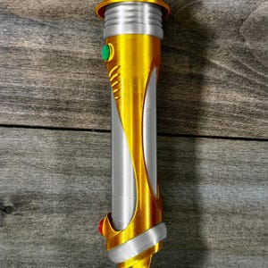 May include: A gold and silver lightsaber prop with a cylindrical body. The hilt features a gold finish with silver accents and a green button. The bottom has a red button. The lightsaber is a prop for cosplay or display.
