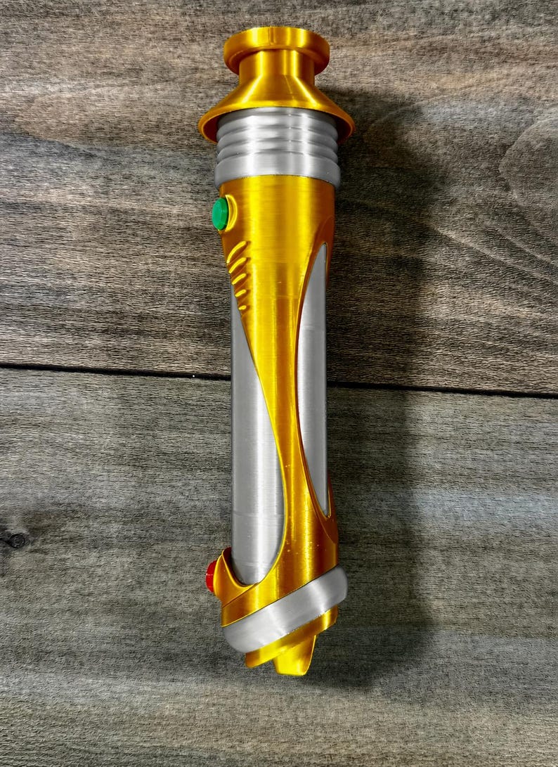 May include: A gold and silver lightsaber prop with a cylindrical body. The hilt features a gold finish with silver accents and a green button. The bottom has a red button. The lightsaber is a prop for cosplay or display.