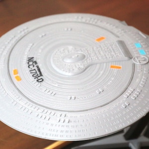 K&ouml;nnte beinhalten: Nahaufnahme eines wei&szlig;en Modells eines Star Trek Raumschiff-Untertassenabschnitts. Der Untertassenabschnitt hat ein detailliertes Design mit einem erh&ouml;hten Mittelteil und einem Ring aus kleineren erh&ouml;hten Abschnitten am Rand. Der Untertassenabschnitt ist mit "NCC-1701-D" beschriftet.