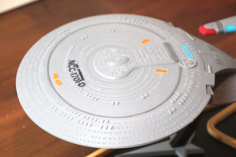 K&ouml;nnte beinhalten: Nahaufnahme eines wei&szlig;en Modells eines Star Trek Raumschiff-Untertassenabschnitts. Der Untertassenabschnitt hat ein detailliertes Design mit einem erh&ouml;hten Mittelteil und einem Ring aus kleineren erh&ouml;hten Abschnitten am Rand. Der Untertassenabschnitt ist mit "NCC-1701-D" beschriftet.