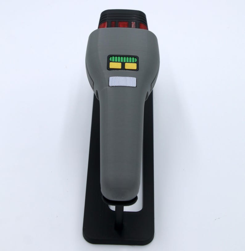 K&ouml;nnte beinhalten: Eine graue Star Trek Phaser-Replik mit einem schwarzen St&auml;nder. Der Phaser hat ein gr&uuml;n-gelbes Display und ein rotes Licht oben.