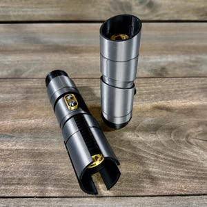May include: Two silver and black lightsaber hilts with gold accents are displayed on a wooden surface. One hilt is lying down, while the other stands upright. The hilts have a cylindrical design with detailed sections.