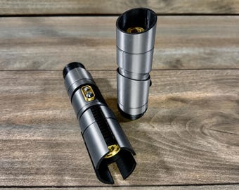 May include: Two silver and black lightsaber hilts with gold accents are displayed on a wooden surface. One hilt is lying down, while the other stands upright. The hilts have a cylindrical design with detailed sections.