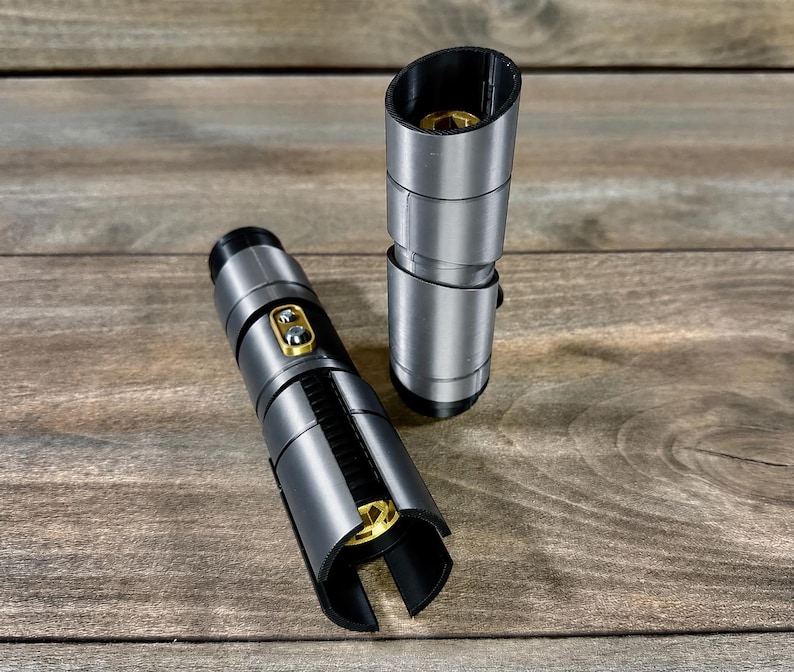 May include: Two silver and black lightsaber hilts with gold accents are displayed on a wooden surface. One hilt is lying down, while the other stands upright. The hilts have a cylindrical design with detailed sections.