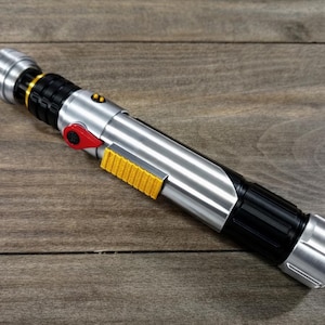 Puede incluir: Un accesorio de sable de luz plateado y negro con un bot&oacute;n rojo y un detalle de agarre amarillo. El accesorio tiene un acabado met&aacute;lico y est&aacute; sobre una superficie de madera. El sable de luz tiene un mango negro y un emisor plateado.