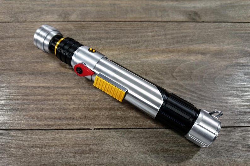 Puede incluir: Un accesorio de sable de luz plateado y negro con un bot&oacute;n rojo y un detalle de agarre amarillo. El accesorio tiene un acabado met&aacute;lico y est&aacute; sobre una superficie de madera. El sable de luz tiene un mango negro y un emisor plateado.