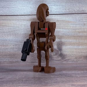 Pode incluir: Uma figura de robô castanho articulada com uma pistola blaster preta. O robô tem uma cabeça arredondada, uma luz vermelha no peito e está em pé. A figura é feita de plástico e é um brinquedo.