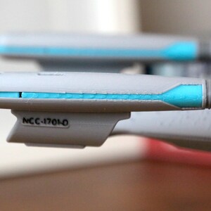 K&ouml;nnte beinhalten: Nahaufnahme eines grauen und wei&szlig;en Raumschiffmodells mit einem blauen Streifen und dem Text "NCC-1701-0" an der Seite.