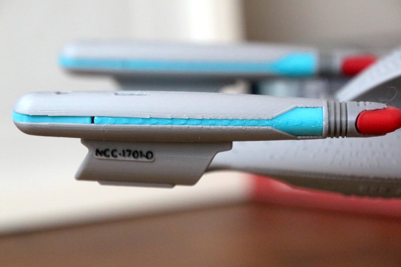 K&ouml;nnte beinhalten: Nahaufnahme eines grauen und wei&szlig;en Raumschiffmodells mit einem blauen Streifen und dem Text "NCC-1701-0" an der Seite.