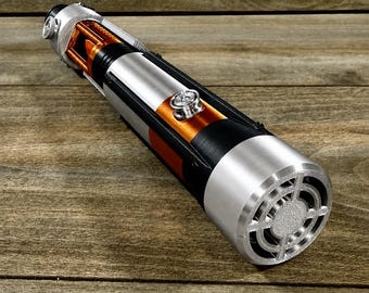 May include: A silver, black, and orange lightsaber prop with intricate detailing. The hilt features a silver finish with orange and black accents. The end has a circular design with a silver finish. The lightsaber is displayed on a wooden surface.