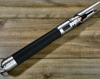 May include: A silver and black lightsaber prop with a textured black grip. The hilt has intricate details and a blade emitter at the top. The lightsaber is displayed on a wooden surface.