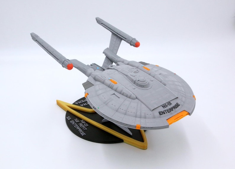 Puede incluir: Maqueta gris de la nave espacial NX-01 Enterprise con detalles en naranja y rojo. El modelo est&aacute; sobre un soporte negro y dorado con el texto "NX-01 Enterprise" y "SS Enterprise". La nave tiene dos largas g&oacute;ndolas de curvatura grises.