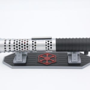 May include: A silver and black cylindrical lightsaber hilt featuring a perforated metal emitter section and a ribbed grip. The hilt rests horizontally on a black and gray display stand adorned with a red Imperial crest emblem in the center.