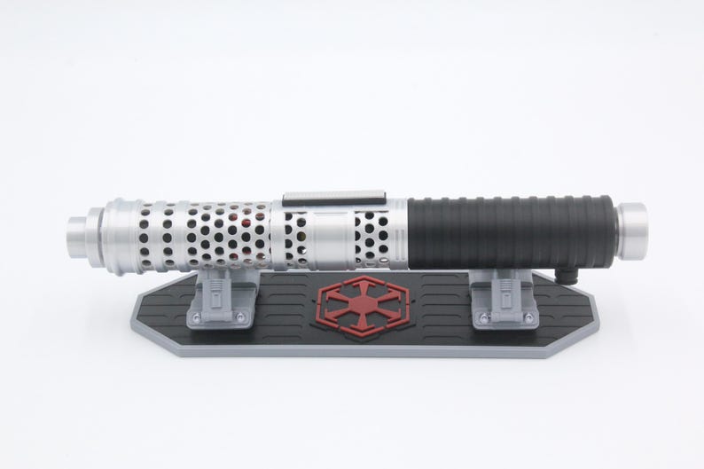 May include: A silver and black cylindrical lightsaber hilt featuring a perforated metal emitter section and a ribbed grip. The hilt rests horizontally on a black and gray display stand adorned with a red Imperial crest emblem in the center.
