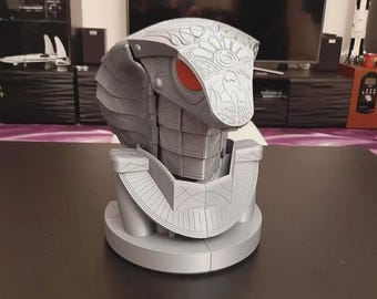 May include: A gray, cobra-shaped sculpture with intricate details, including a detailed head and body. The cobra has red eyes and is set on a circular base. The sculpture is on a dark surface.