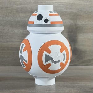 May include: A white BB-8 droid figure with orange and grey accents. The spherical body features large orange and grey circular designs. The head has orange stripes and black and grey details. The figure is a collectible item.